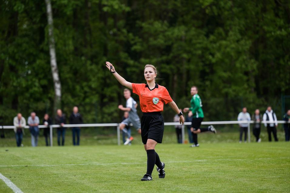 Schiedsrichterin mit orangefarbener Uniform zeigt beim Fußballspiel ein Zeichen auf dem grünen Spielfeld.